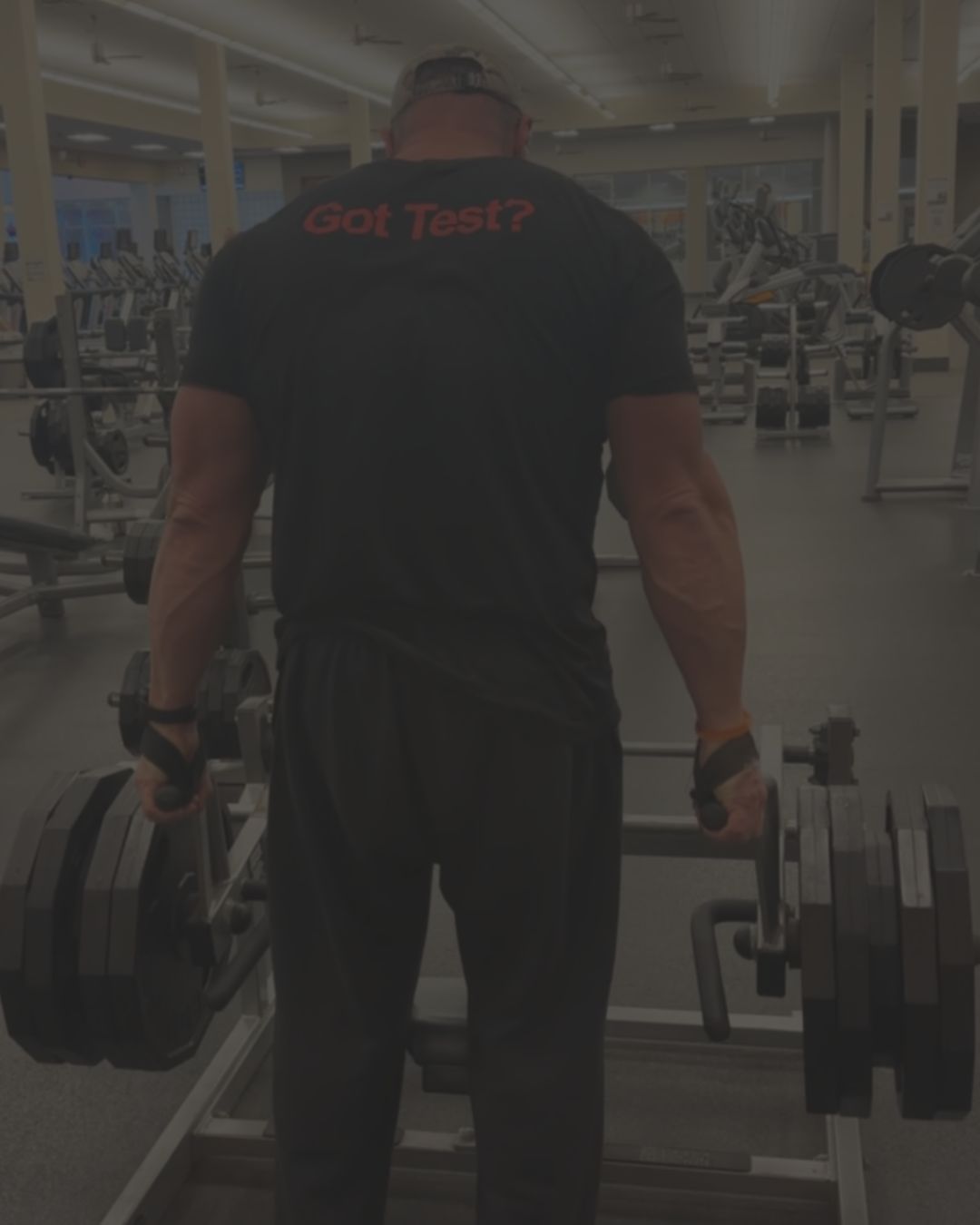 A man viewed from behind in a gym, wearing a black t-shirt with 'Got Test?' printed in red on the back, holding a barbell on a bench.
