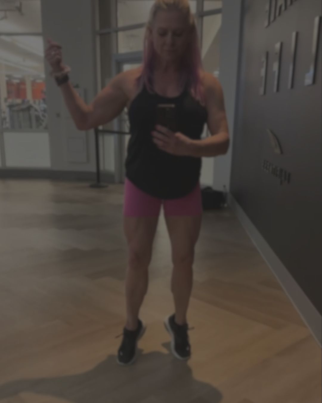 A woman in a gym wearing a black tank top and pink athletic shorts, standing on a wooden floor.