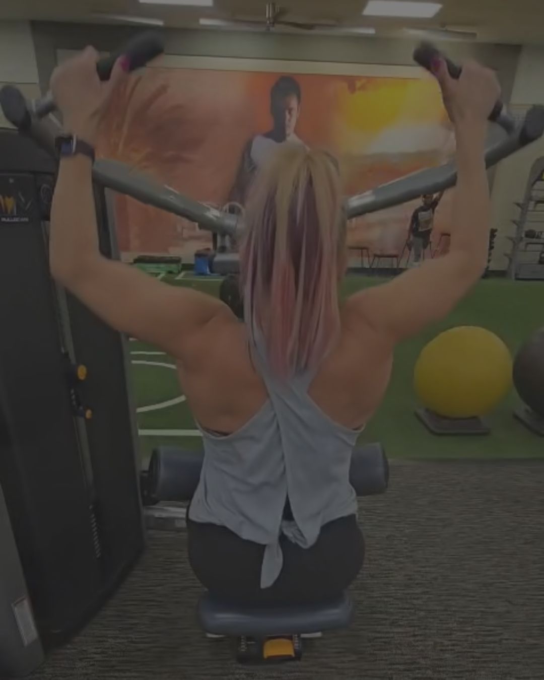 A woman with pink streaked hair is seen from behind, using a chest-supported seated cable machine in a gym setting.