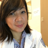 A close-up portrait of a woman wearing a white medical lab coat over a blue and white striped collared shirt, with a warm indoor hallway background.