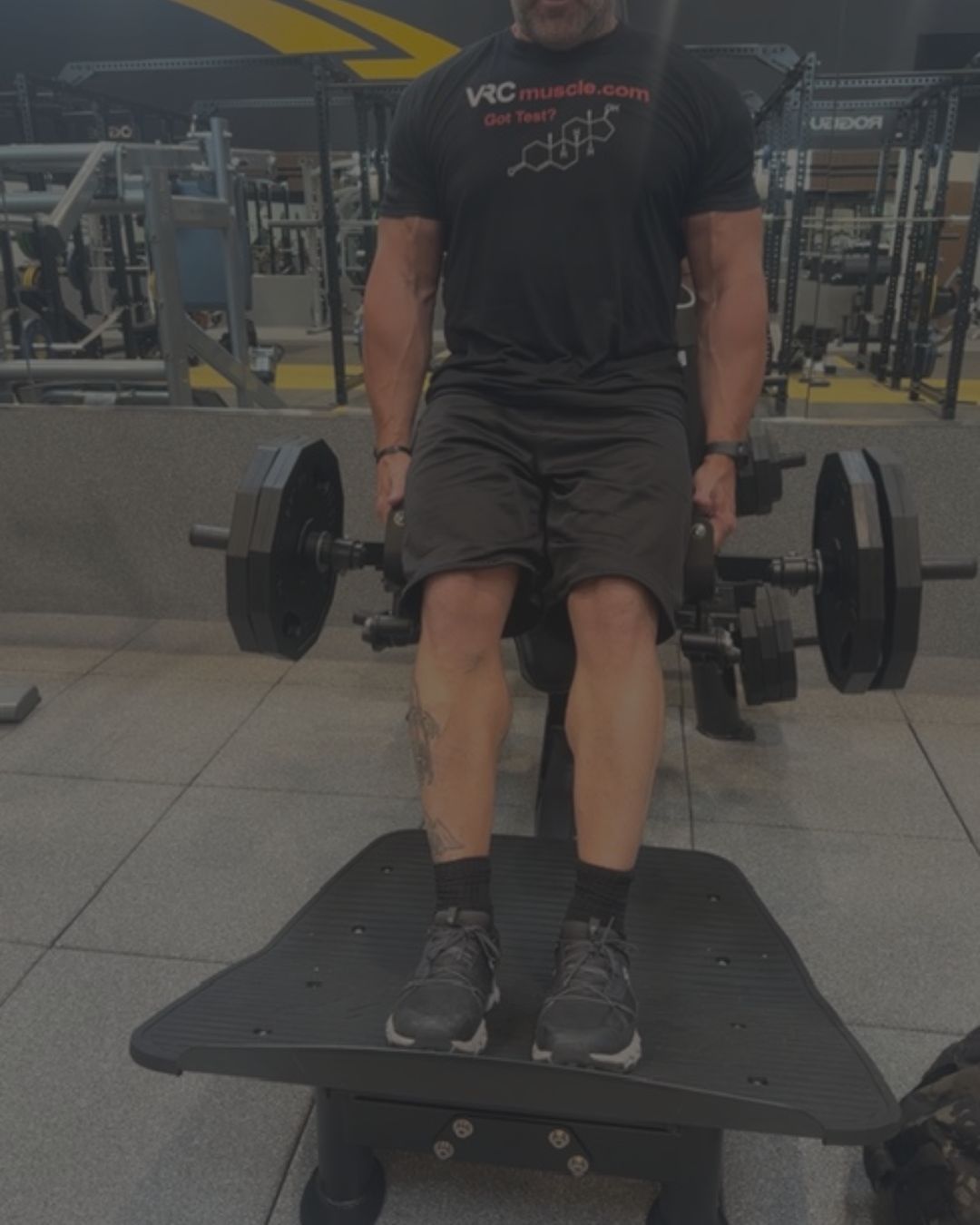 A man in a gym wearing a black t-shirt and shorts, standing on a black platform while holding a barbell with weight plates in each hand.