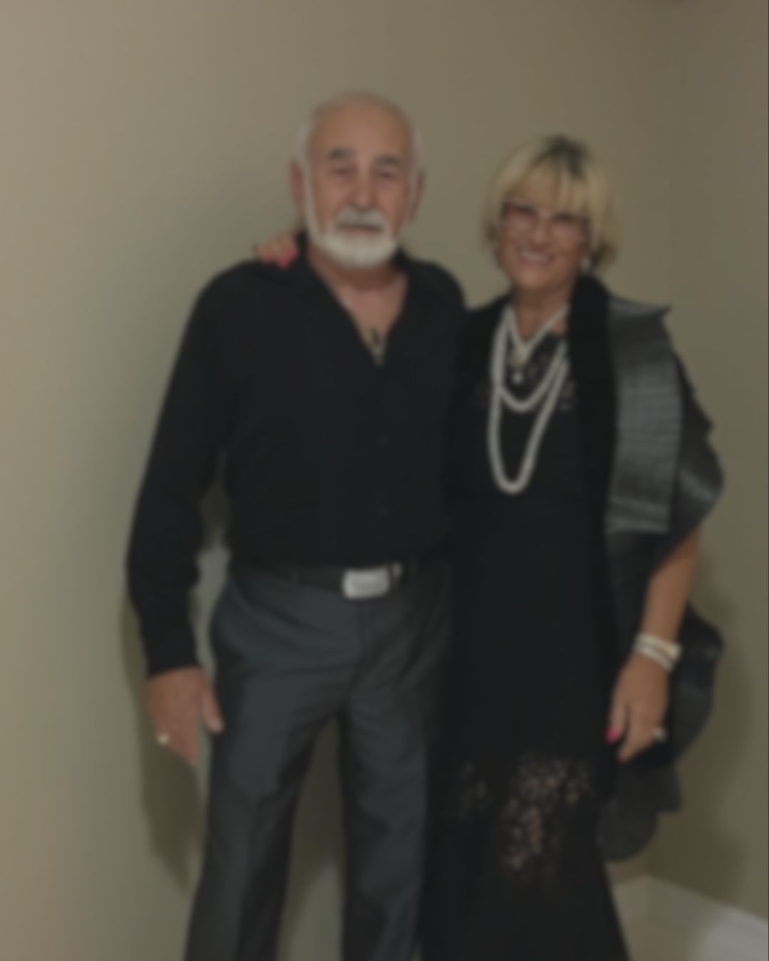 An older man with a white beard and mustache wearing a black long-sleeve shirt and gray trousers, standing next to an older woman with short blonde hair wearing a black dress, a gray shawl, and multiple white pearl necklaces.