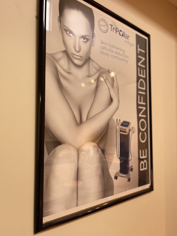 A framed TriPolar skin tightening poster hanging on a beige wall. The poster features a black-and-white photo of a seated woman with small yellow light dots overlaid, alongside a small depiction of a TriPolar device and the text 'BE CONFIDENT' and 'TriPolar'.
