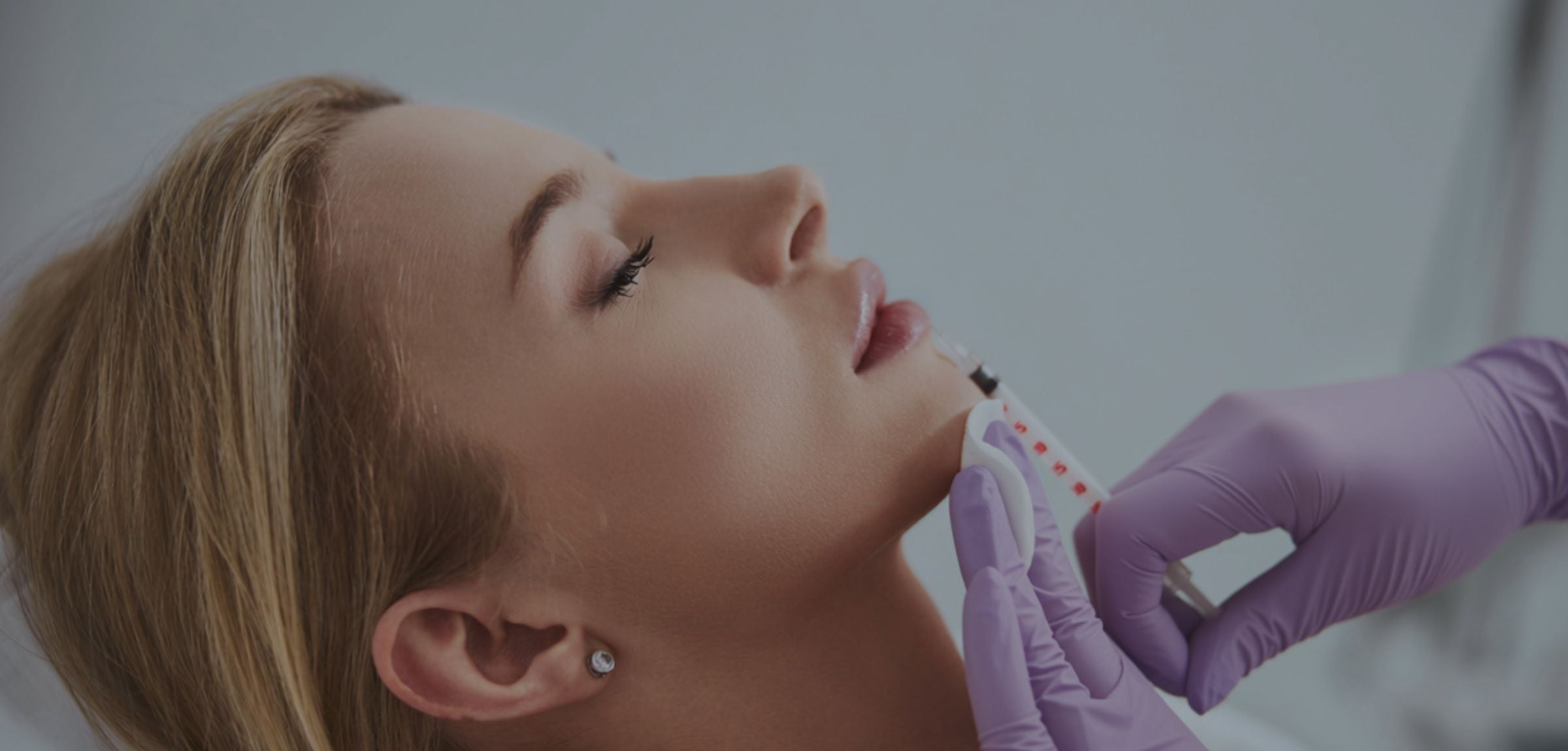 A close-up of a woman receiving a lip blushing tattoo or cosmetic lip treatment, with a gloved hand holding a small applicator near her lips. The woman has blonde hair and a small stud earring, and the background is a soft, out-of-focus light gray. Vitality Rejuvenation offers aesthetics such as lip filler, dermal fillers, and more from their Cherry Hill office. 