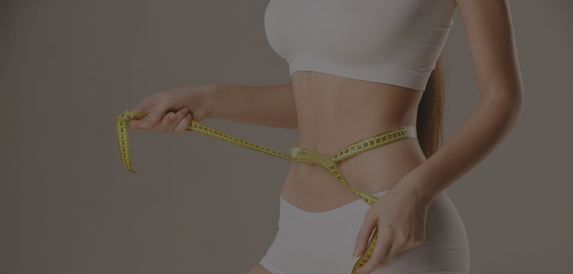 A woman in a white sports bra and shorts measuring her waist with a yellow tape measure against a plain taupe background.